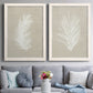 Set of two white barnwood framed canvas prints depicting paired minimalist tropical botanical prints with feathery leaves, warm taupe grounds, and simple high-contrast shapes above a sofa in a contemporary living room.