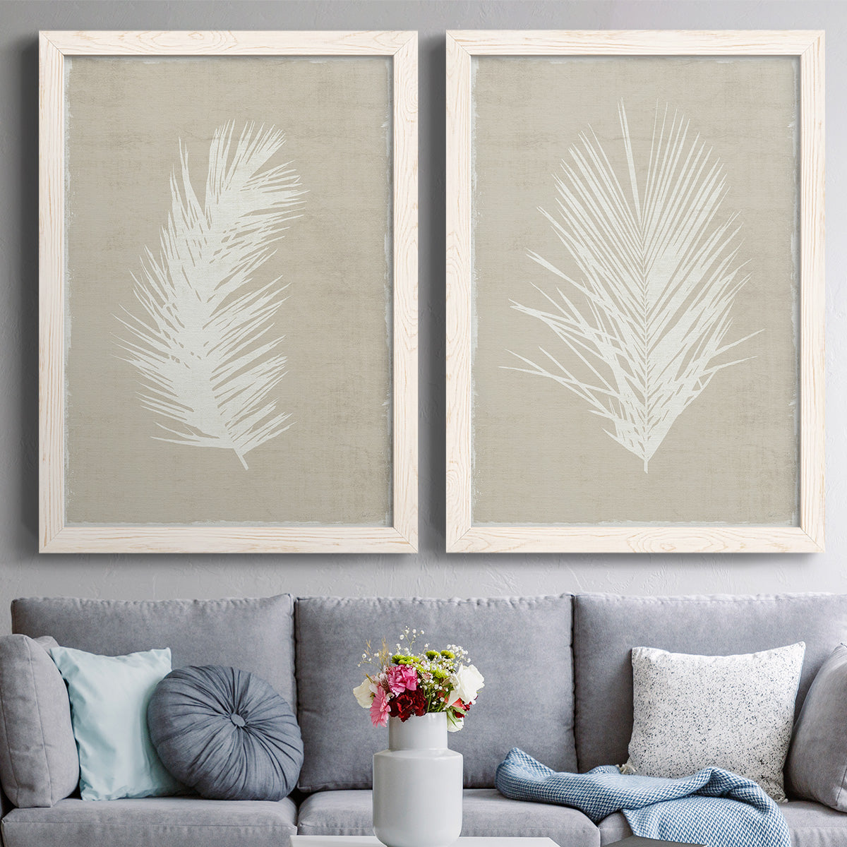 Set of two white barnwood framed canvas prints depicting paired minimalist tropical botanical prints with feathery leaves, warm taupe grounds, and simple high-contrast shapes above a sofa in a contemporary living room.