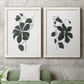 Set of two white barnwood framed canvas prints depicting a coordinated art pair of black leafy vine silhouettes rendered as minimalist botanical prints above a sofa in a modern living room.