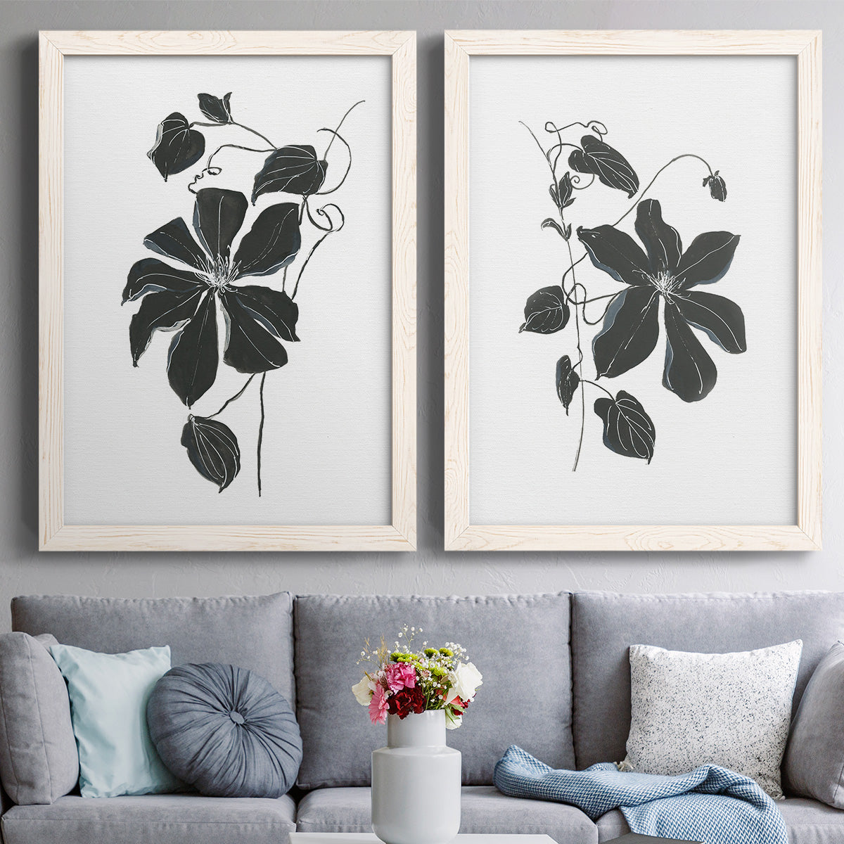 Set of two white barnwood framed canvas prints depicting paired minimalist botanical prints with bold dark leaves, curving stems, and clean white grounds above a sofa in a contemporary living room.