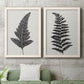 Set of two white barnwood framed canvas prints depicting a coordinated art pair of black fern leaf silhouettes rendered as minimalist botanical prints above a sofa in a modern living room.