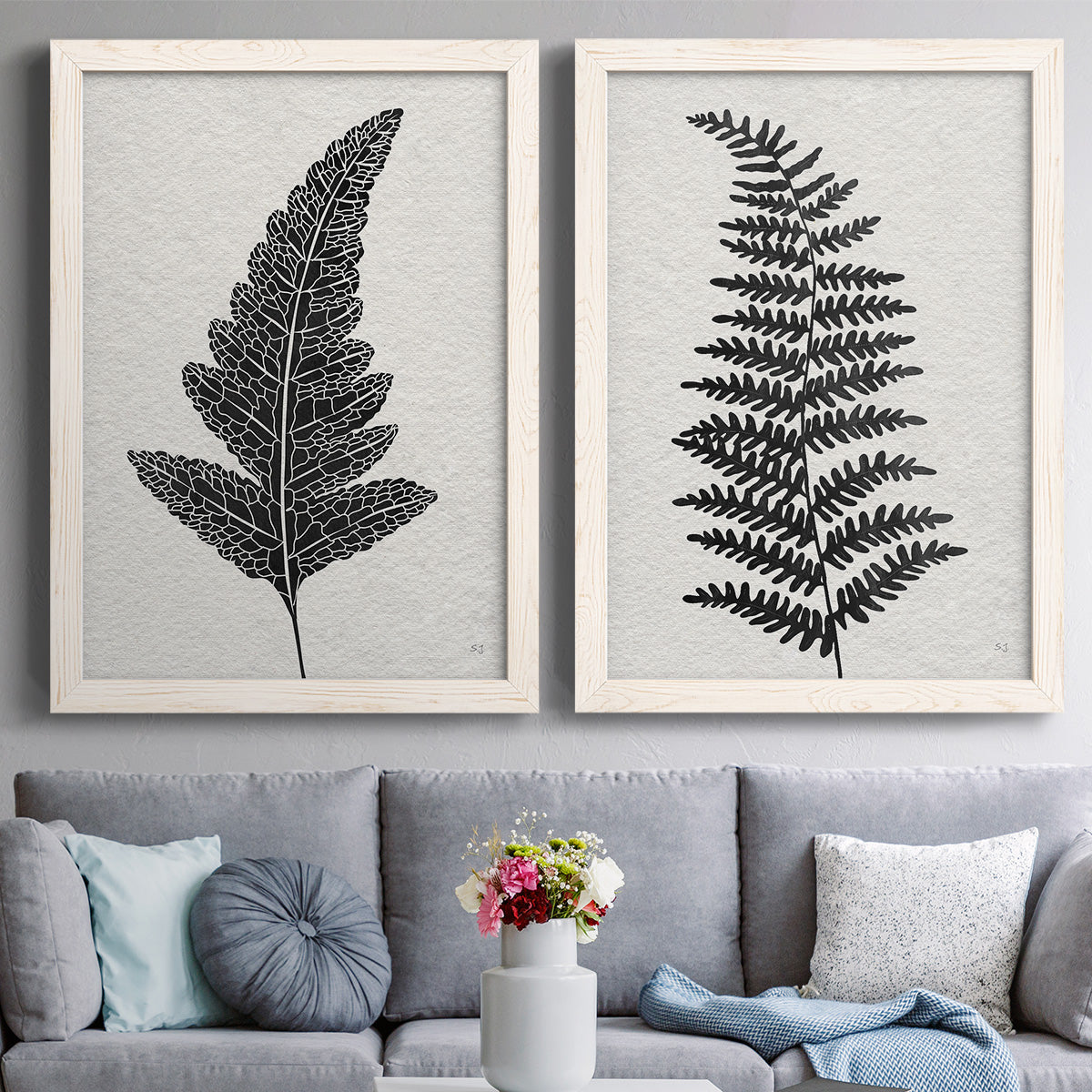 Set of two white barnwood framed canvas prints depicting paired minimalist botanical prints with detailed fronds, high-contrast shapes, and lightly textured cream grounds above a sofa in a contemporary living room.
