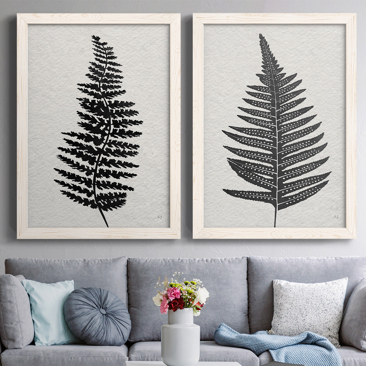Set of two white barnwood framed canvas prints depicting paired minimalist botanical prints with detailed fronds, high-contrast shapes, and lightly textured cream grounds above a sofa in a contemporary living room.