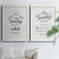 Set of two white barnwood framed canvas prints depicting hand-lettered phrases, socks hangers and laundry symbols on linen texture arranged across two complementary compositions above a sofa in a bright living space.