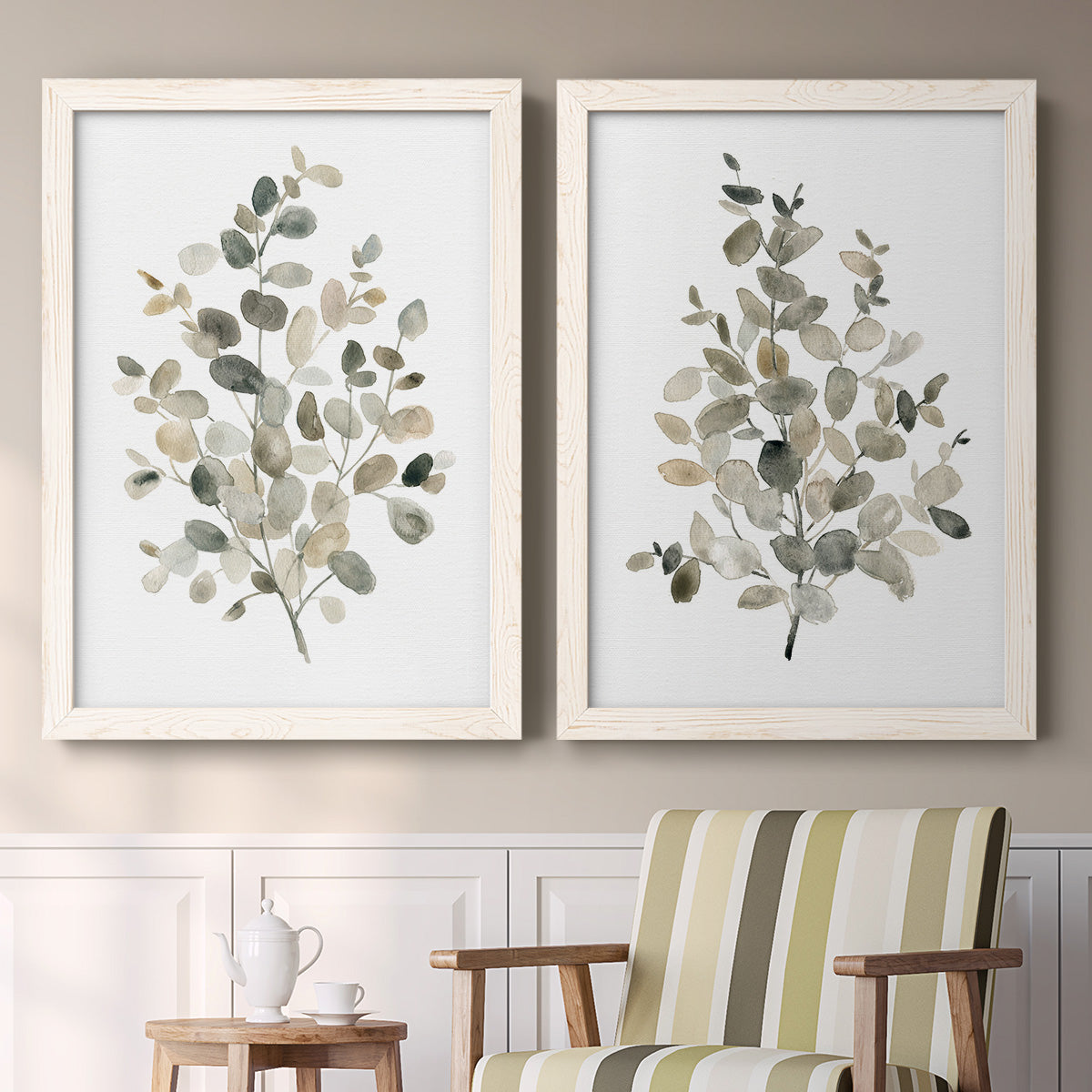 Set of two white barnwood framed canvas prints depicting two minimalist watercolor botanical studies featuring delicate eucalyptus branches with rounded leaves above a sideboard in a cozy sitting room.