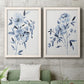 Set of two white barnwood framed canvas prints depicting a coordinated art pair of loose blue flowers and leafy branches rendered as expressive watercolor botanical paintings above a sofa in a modern living room.