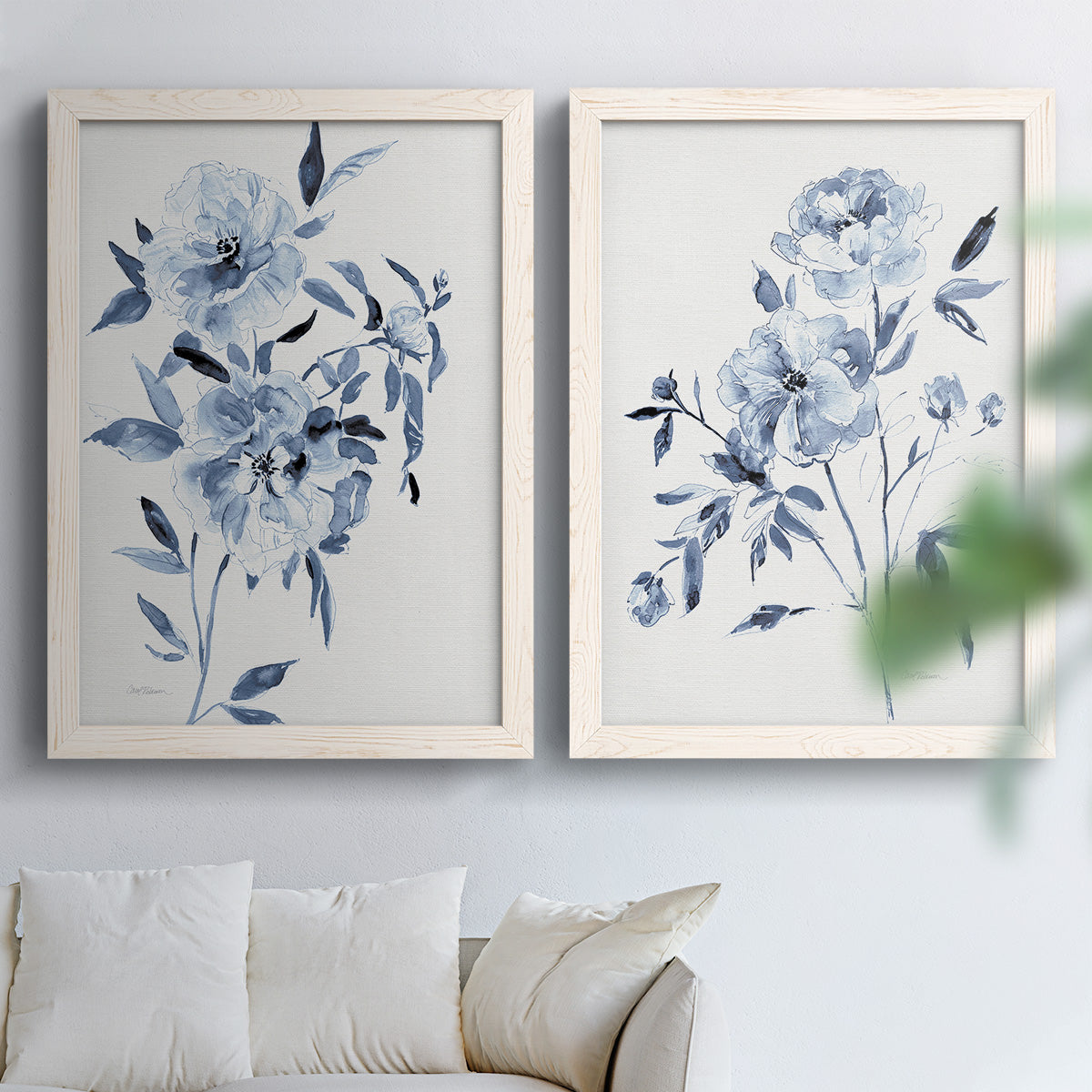Set of two white barnwood framed canvas prints depicting indigo blossoms, gestural blue leaves, and airy white space arranged across two complementary compositions above a sofa in a bright living space.