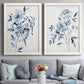 Set of two white barnwood framed canvas prints depicting paired expressive watercolor botanical paintings with indigo blossoms, gestural blue leaves, and airy white space above a sofa in a contemporary living room.