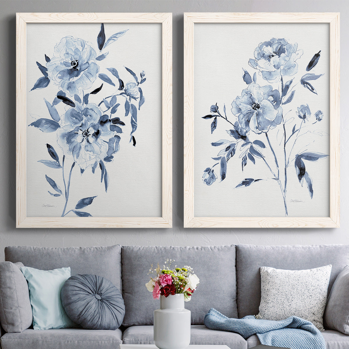 Set of two white barnwood framed canvas prints depicting paired expressive watercolor botanical paintings with indigo blossoms, gestural blue leaves, and airy white space above a sofa in a contemporary living room.
