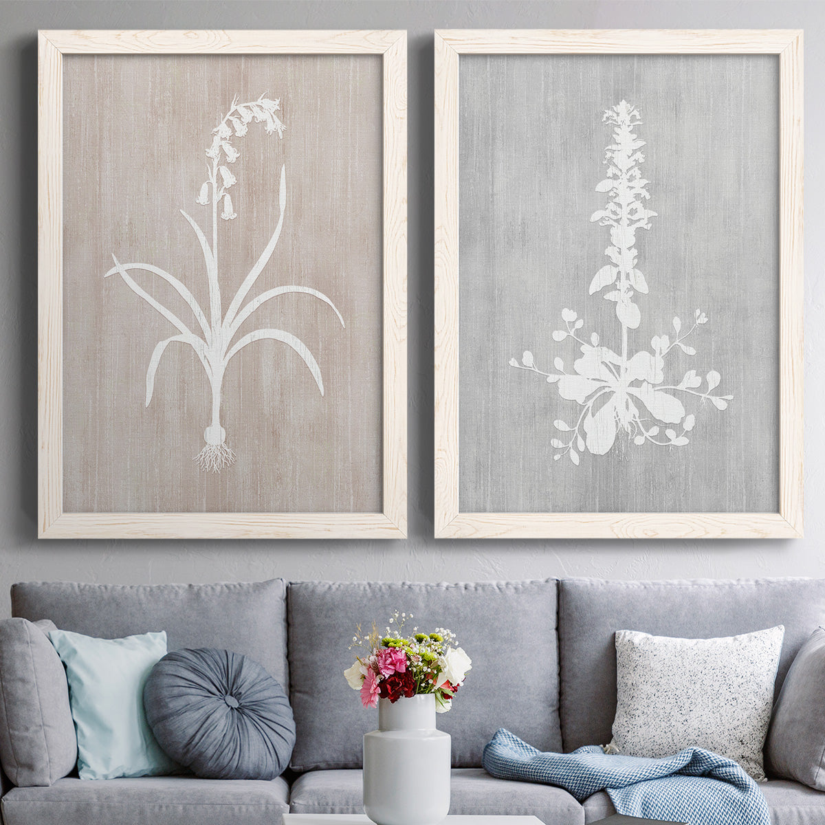 Set of two white barnwood framed canvas prints depicting paired rustic minimalist botanical prints with delicate stems, leaves, and blossoms against gray taupe and pale blue panels above a sofa in a contemporary living room.