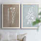Set of two white barnwood framed canvas prints depicting delicate stems, leaves, and blossoms against gray taupe and pale blue panels arranged across two complementary compositions above a sofa in a bright living space.