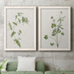 Set of two white barnwood framed canvas prints depicting a coordinated art pair of delicate wildflower and leafy stems rendered as rustic minimalist botanical studies above a sofa in a modern living room.