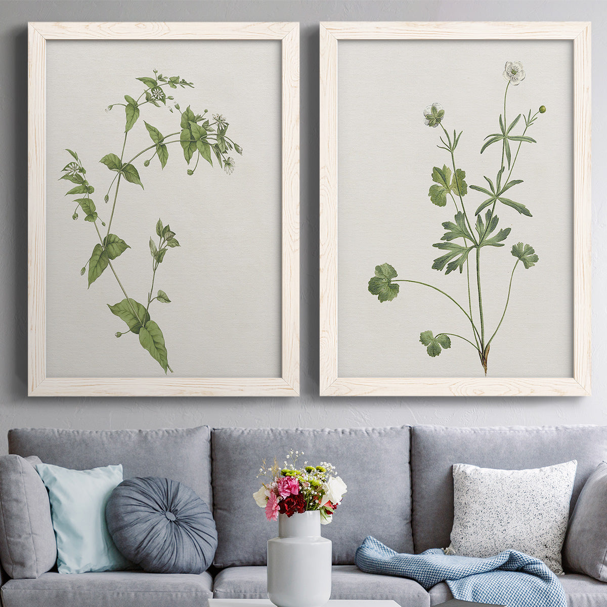 Set of two white barnwood framed canvas prints depicting paired rustic minimalist botanical studies with fine green branches, sparse blue or white blossoms, and cream grounds above a sofa in a contemporary living room.