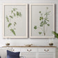 Set of two white barnwood framed canvas prints depicting a coordinated art pair of delicate wildflower and leafy stems rendered as rustic minimalist botanical studies above a dining room sideboard.
