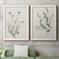 Set of two white barnwood framed canvas prints depicting a coordinated art pair of delicate wildflower and leafy stems rendered as rustic minimalist botanical studies above a sofa in a modern living room.