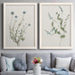 Set of two white barnwood framed canvas prints depicting paired rustic minimalist botanical studies with fine green branches, sparse blue or white blossoms, and cream grounds above a sofa in a contemporary living room.