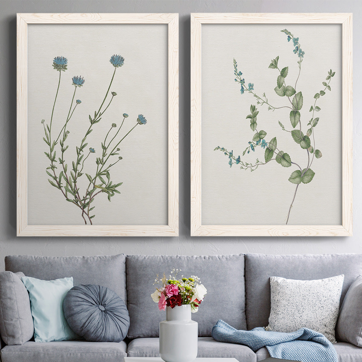 Set of two white barnwood framed canvas prints depicting paired rustic minimalist botanical studies with fine green branches, sparse blue or white blossoms, and cream grounds above a sofa in a contemporary living room.
