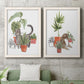 Set of two white barnwood framed canvas prints depicting a coordinated art pair of cats tucked among collections of potted houseplants rendered as whimsical botanical animal illustrations above a sofa in a modern living room.