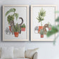 Set of two white barnwood framed canvas prints depicting curious felines, terracotta pots, succulents, and leafy stems arranged across two complementary compositions above a sofa in a bright living space.