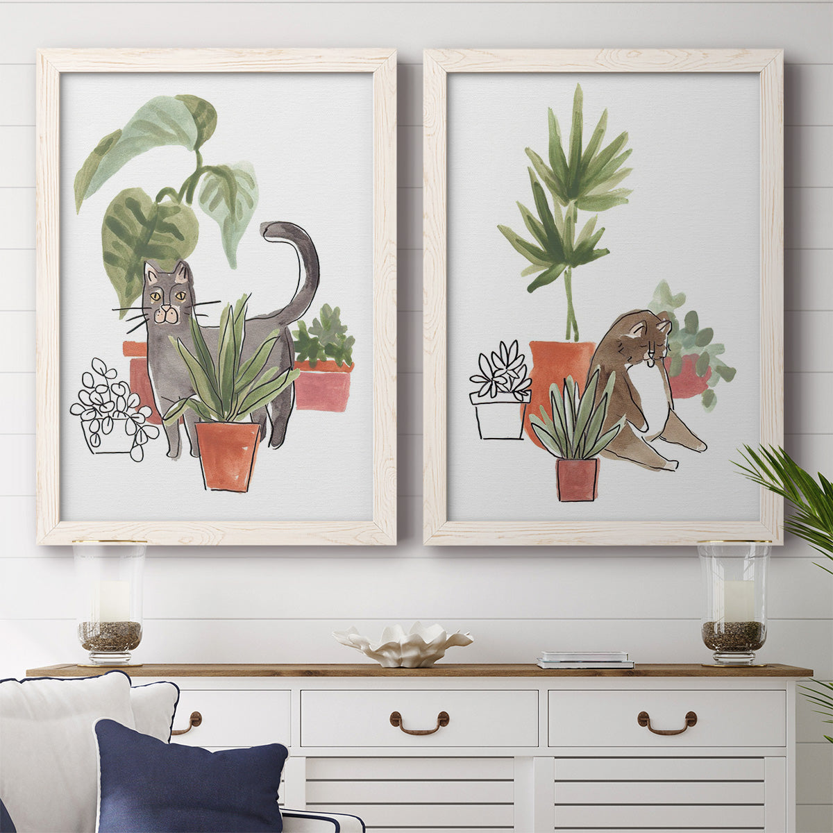 Set of two white barnwood framed canvas prints depicting a coordinated art pair of cats tucked among collections of potted houseplants rendered as whimsical botanical animal illustrations above a dining room sideboard.