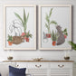 Set of two white barnwood framed canvas prints depicting a coordinated art pair of cats tucked among collections of potted houseplants rendered as whimsical botanical animal illustrations above a dining room sideboard.