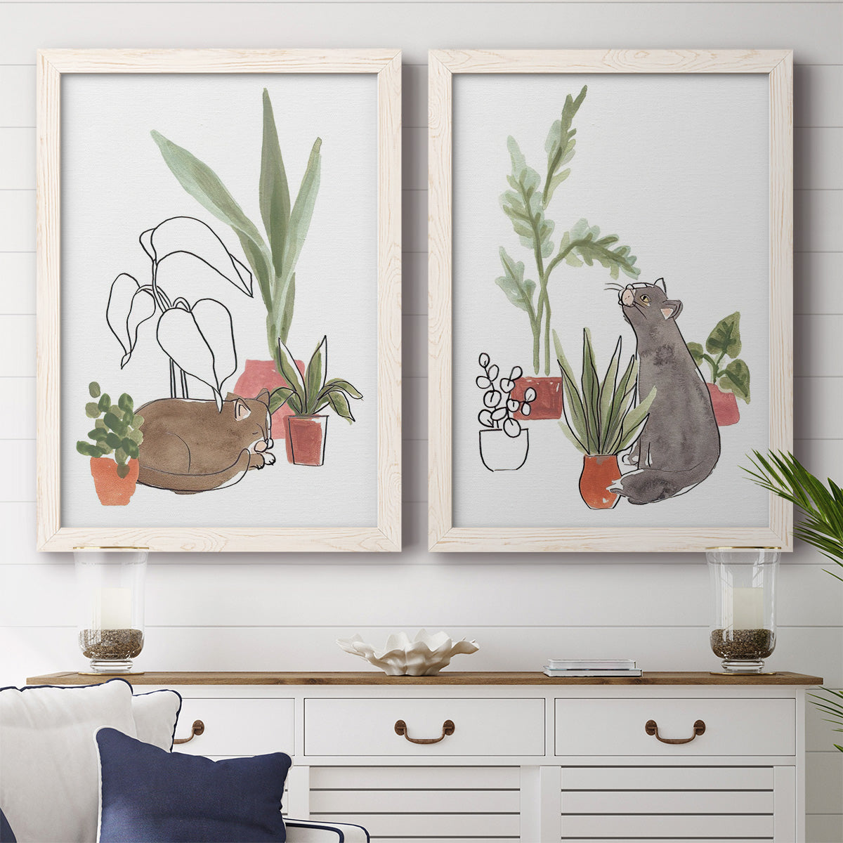 Set of two white barnwood framed canvas prints depicting a coordinated art pair of cats tucked among collections of potted houseplants rendered as whimsical botanical animal illustrations above a dining room sideboard.
