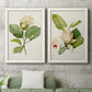 Set of two white barnwood framed canvas prints depicting a coordinated art pair of white magnolia blossoms and glossy green leaves rendered as traditional botanical paintings above a sofa in a modern living room.