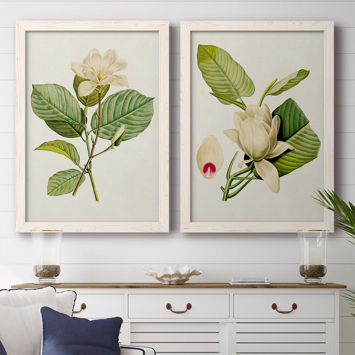 Set of two white barnwood framed canvas prints depicting a coordinated art pair of white magnolia blossoms and glossy green leaves rendered as traditional botanical paintings above a dining room sideboard.