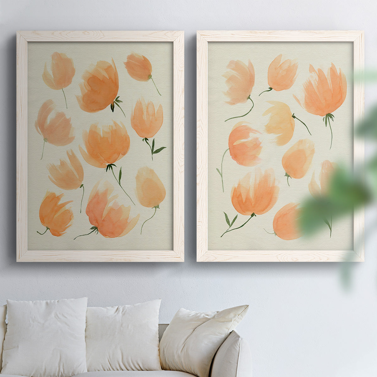 Set of two white barnwood framed canvas prints depicting soft petals, thin black stems, and warm cream backgrounds arranged across two complementary compositions above a sofa in a bright living space.