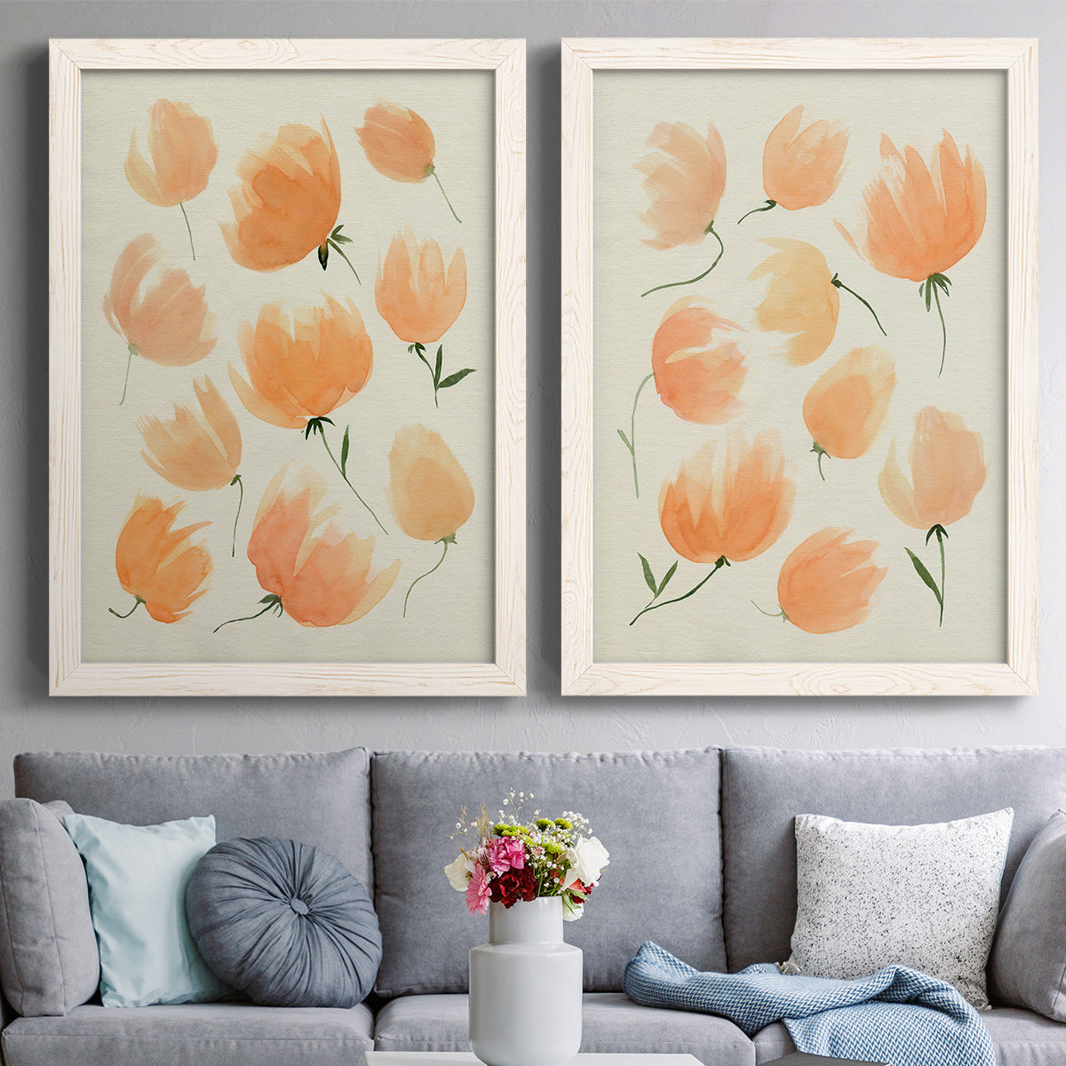 Set of two white barnwood framed canvas prints depicting paired airy watercolor floral paintings with soft petals, thin black stems, and warm cream backgrounds above a sofa in a contemporary living room.