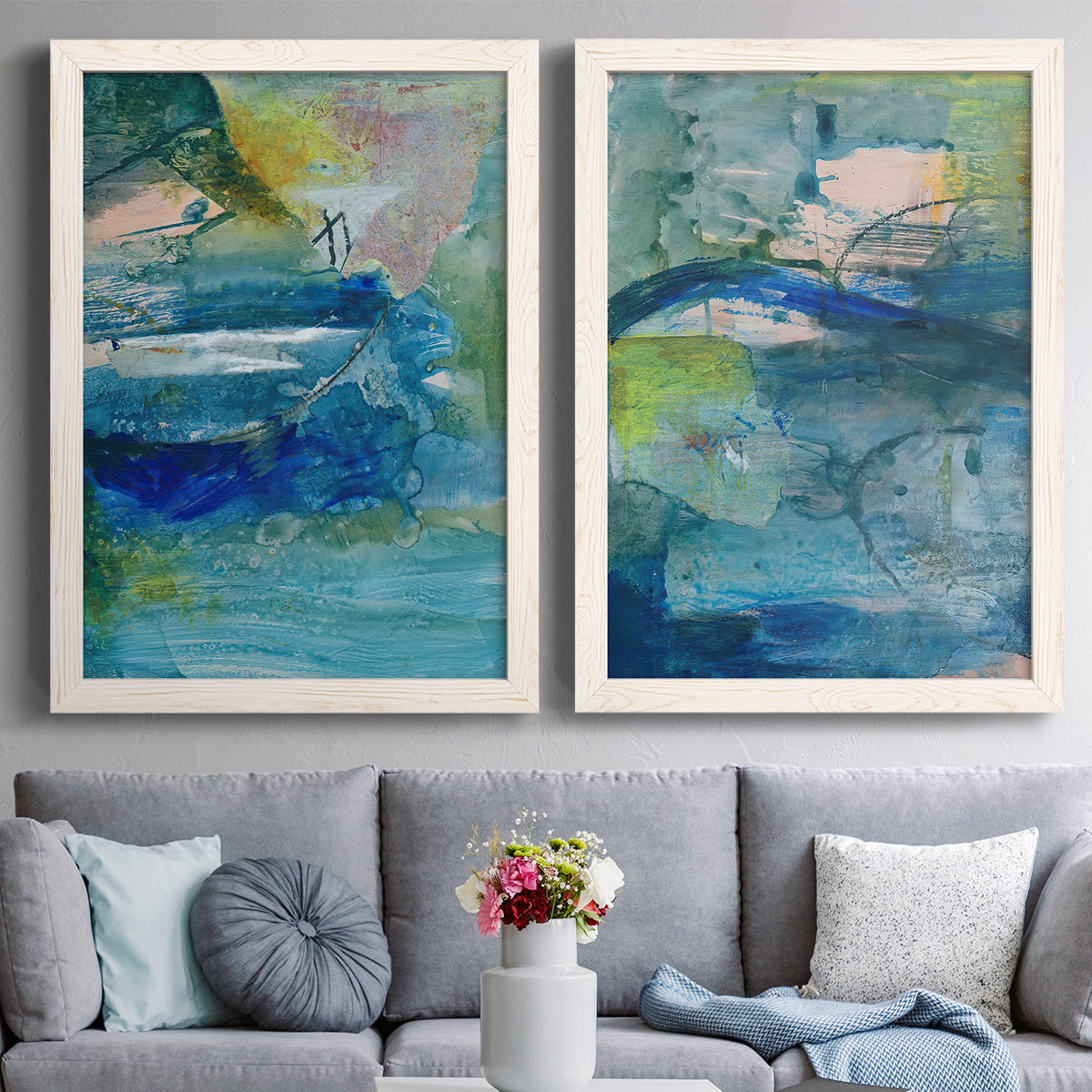 Set of two white barnwood framed canvas prints depicting paired energetic mixed-media abstracts with broad strokes, scratched lines, and luminous color passages above a sofa in a contemporary living room.