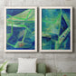 Set of two white barnwood framed canvas prints depicting a coordinated art pair of interlocking blue and green angular forms rendered as textured geometric abstracts above a sofa in a modern living room.