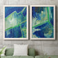 Set of two white barnwood framed canvas prints depicting a coordinated art pair of interlocking blue and green angular forms rendered as textured geometric abstracts above a sofa in a modern living room.