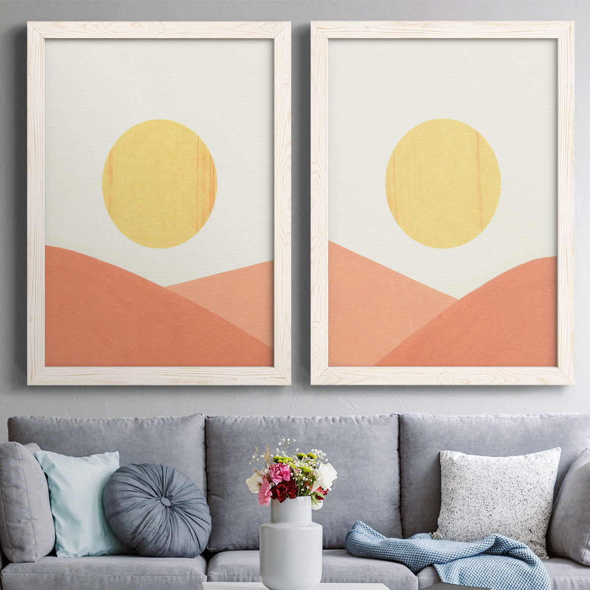 Set of two white barnwood framed canvas prints depicting paired minimalist boho landscapes with flat semicircles, warm peach slopes, and clean cream skies above a sofa in a contemporary living room.