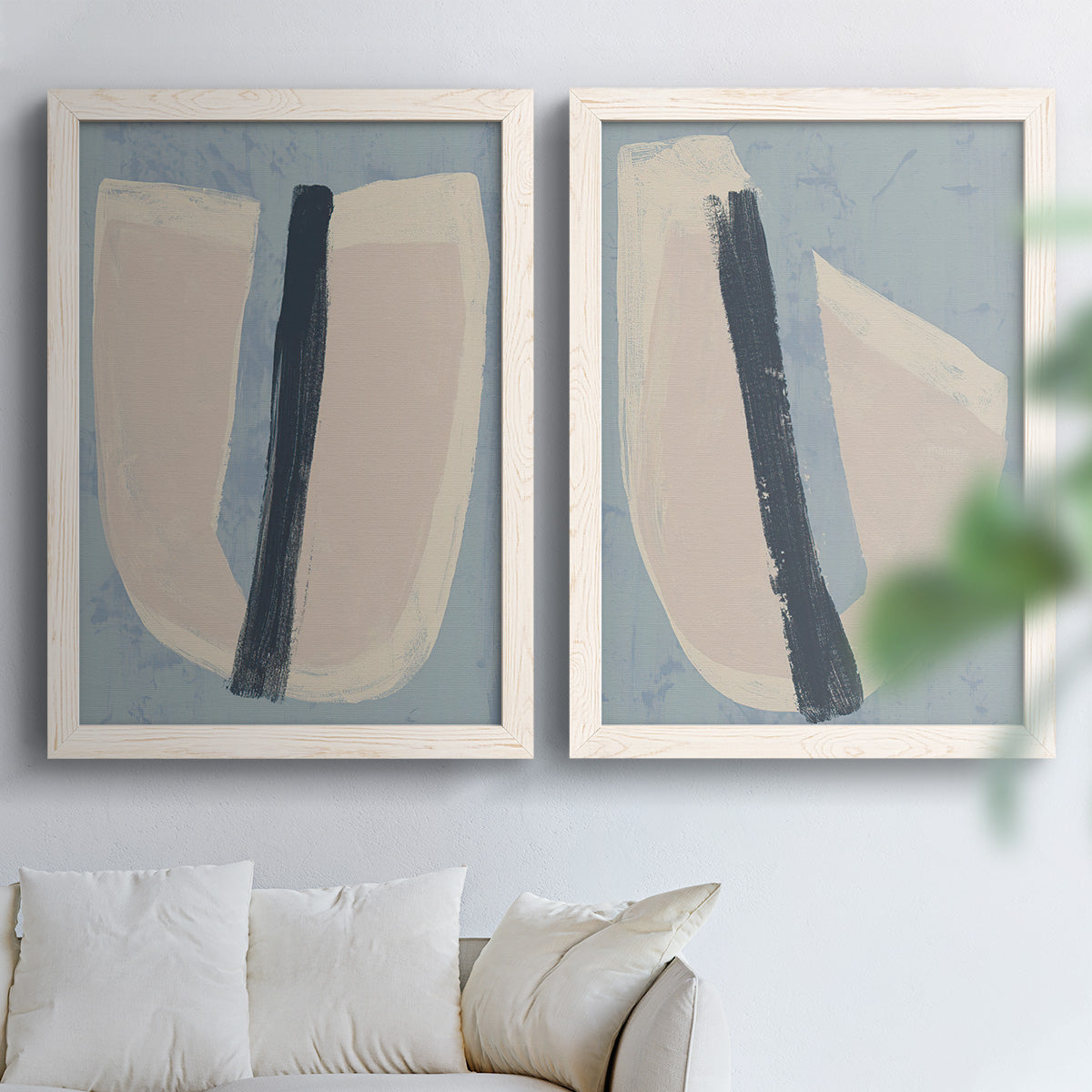 Set of two white barnwood framed canvas prints depicting muted blue green beige fields, torn-edge forms, and bold dark strokes arranged across two complementary compositions above a sofa in a bright living space.