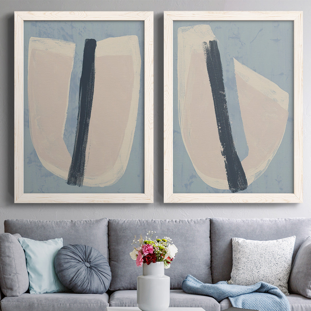 Set of two white barnwood framed canvas prints depicting paired minimalist collage-style abstracts with muted blue green beige fields, torn-edge forms, and bold dark strokes above a sofa in a contemporary living room.