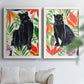 Set of two white barnwood framed canvas prints depicting dark feline figures, broad green leaves, and bright red and orange flowers arranged across two complementary compositions above a sofa in a bright living space.