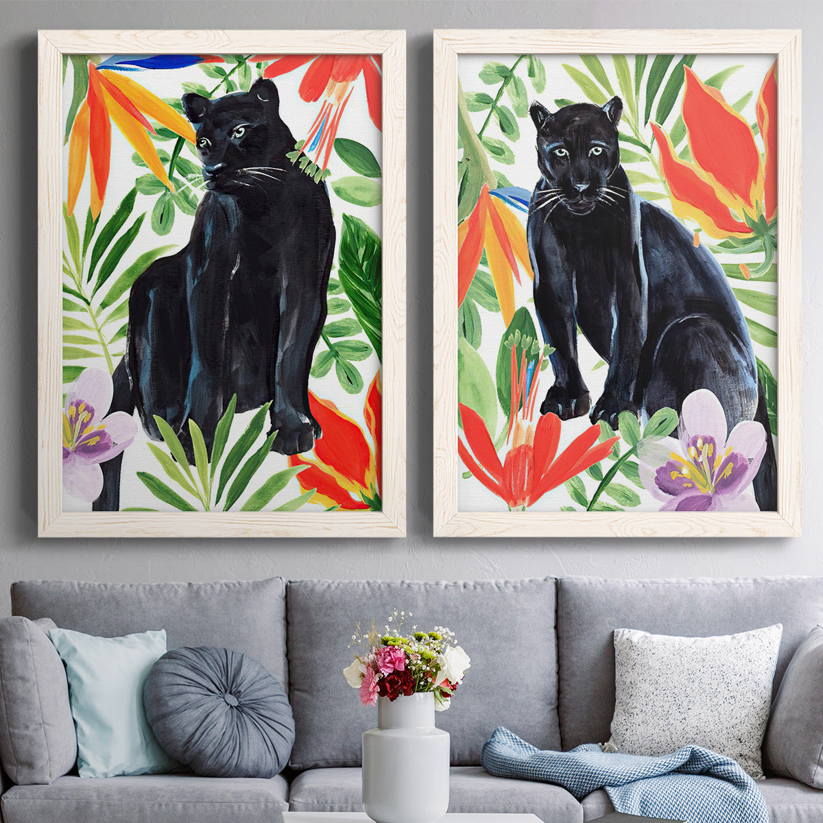 Set of two white barnwood framed canvas prints depicting paired vibrant watercolor wildlife illustrations with dark feline figures, broad green leaves, and bright red and orange flowers above a sofa in a contemporary living room.