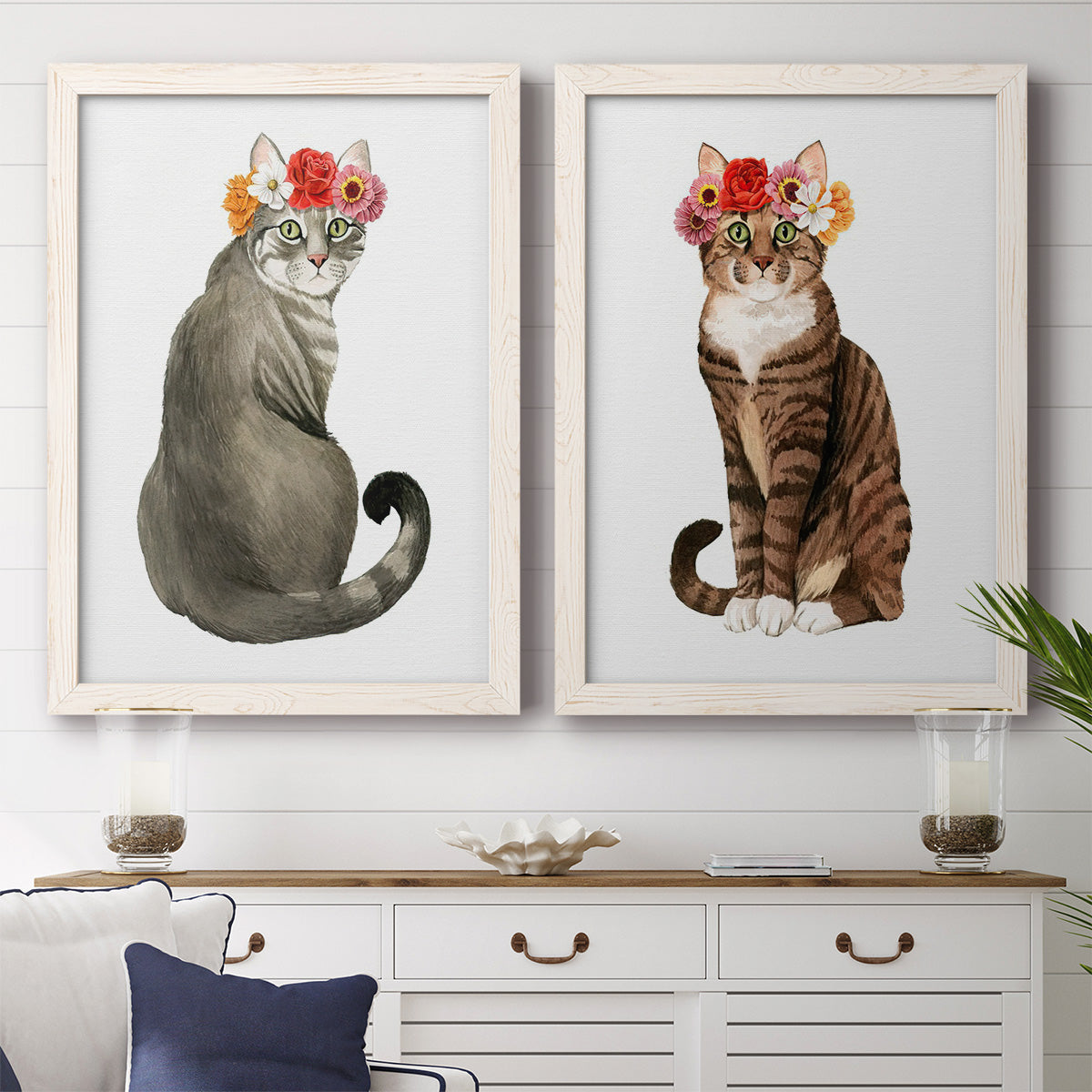 Set of two white barnwood framed canvas prints depicting a coordinated art pair of seated cats wearing colorful flower crowns rendered as whimsical animal portraits above a dining room sideboard.