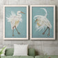 Set of two white barnwood framed canvas prints depicting a coordinated art pair of white herons displaying long windswept feathers rendered as decorative bird illustrations above a sofa in a modern living room.