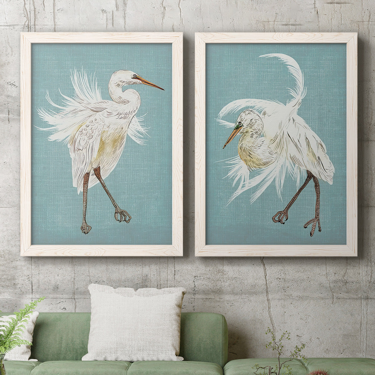 Set of two white barnwood framed canvas prints depicting a coordinated art pair of white herons displaying long windswept feathers rendered as decorative bird illustrations above a sofa in a modern living room.