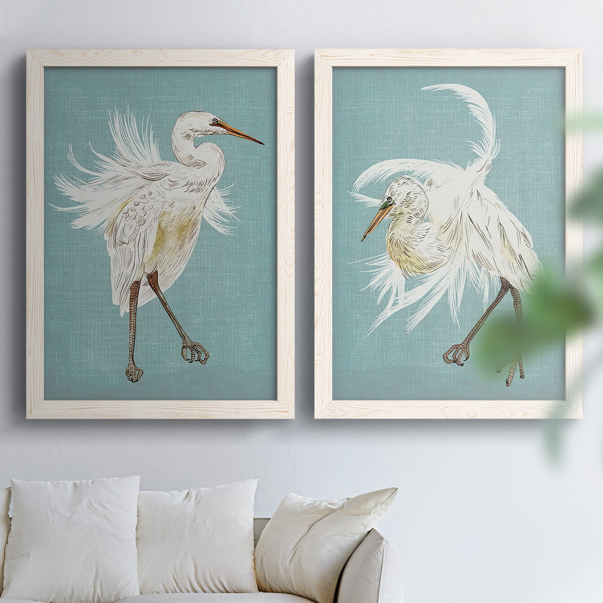 Set of two white barnwood framed canvas prints depicting elegant wading birds, sweeping plumage, and solid olive blue or ochre grounds arranged across two complementary compositions above a sofa in a bright living space.