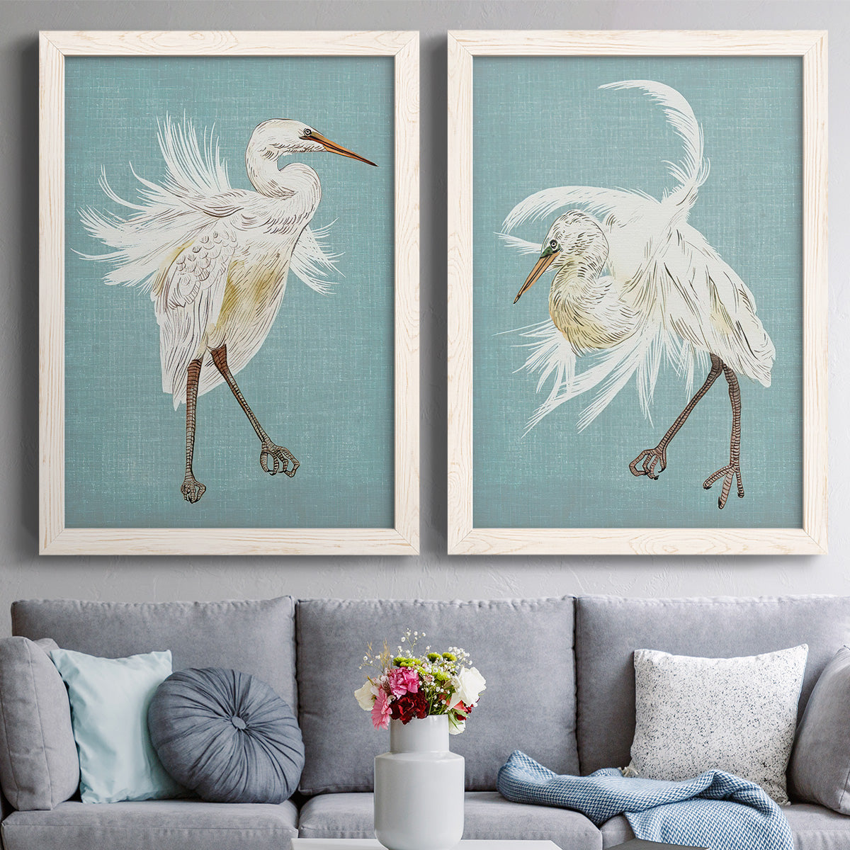 Set of two white barnwood framed canvas prints depicting paired decorative bird illustrations with elegant wading birds, sweeping plumage, and solid olive blue or ochre grounds above a sofa in a contemporary living room.