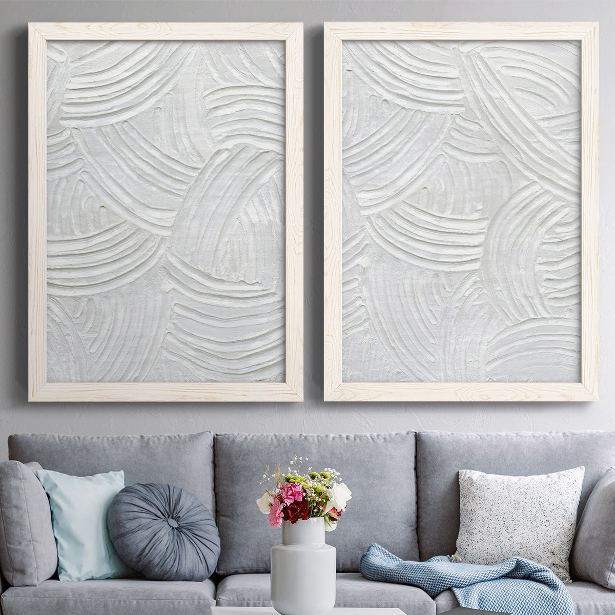 Set of two white barnwood framed canvas prints depicting paired textural monochrome abstracts with raised sweeping grooves, soft shadows, and pale stone-like surfaces above a sofa in a contemporary living room.