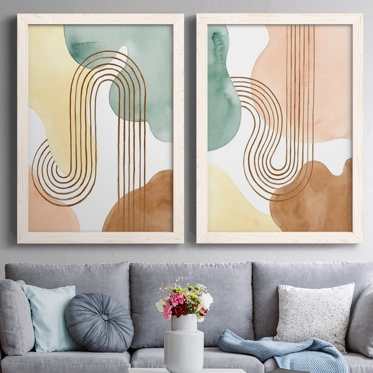 Set of two white barnwood framed canvas prints depicting paired folk-inspired botanical patterns with stylized bouquets, blue block-print forms, and warm white grounds above a sofa in a contemporary living room.