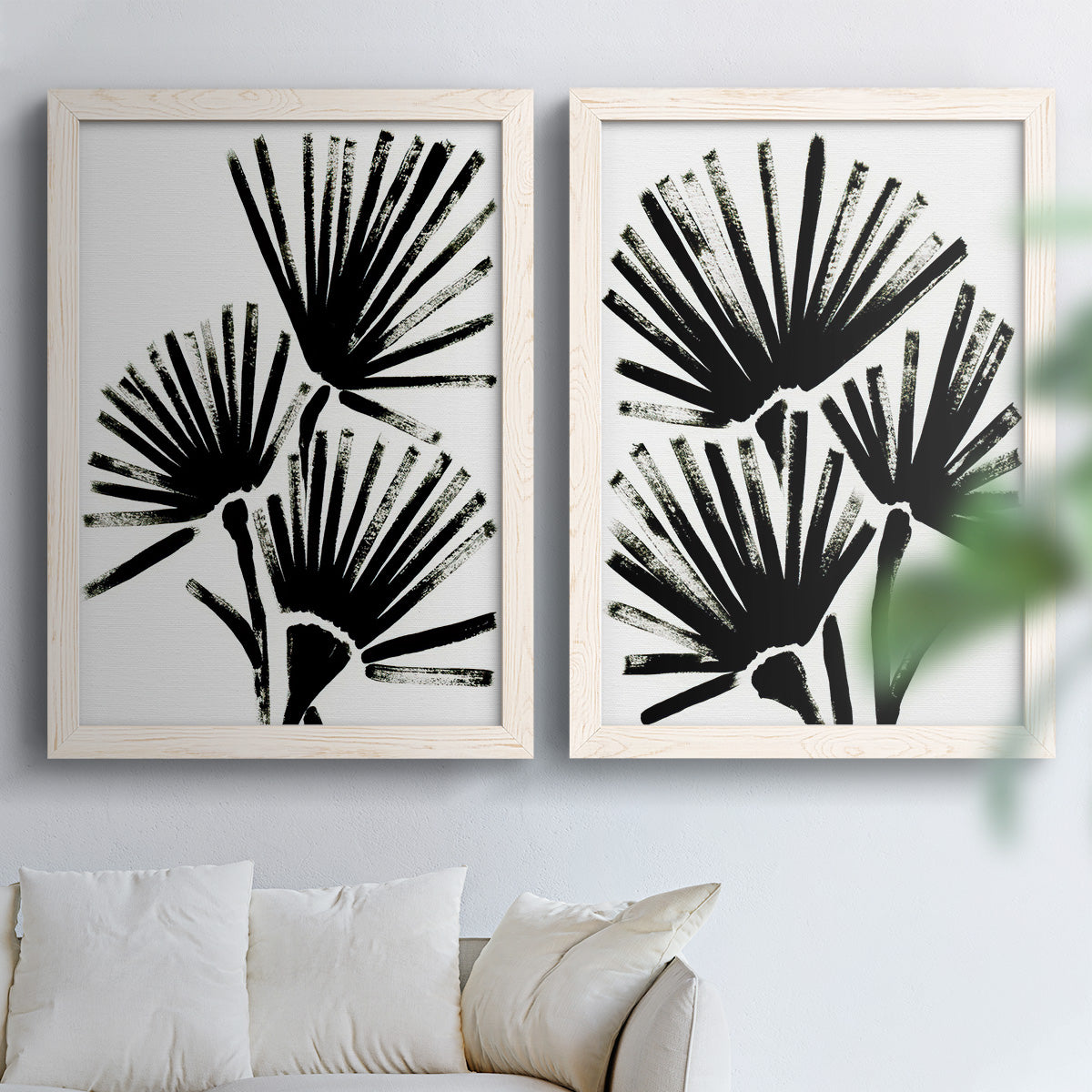 Set of two white barnwood framed canvas prints depicting radiating brush marks, repeated leaf-like fans, and white grounds arranged across two complementary compositions above a sofa in a bright living space.