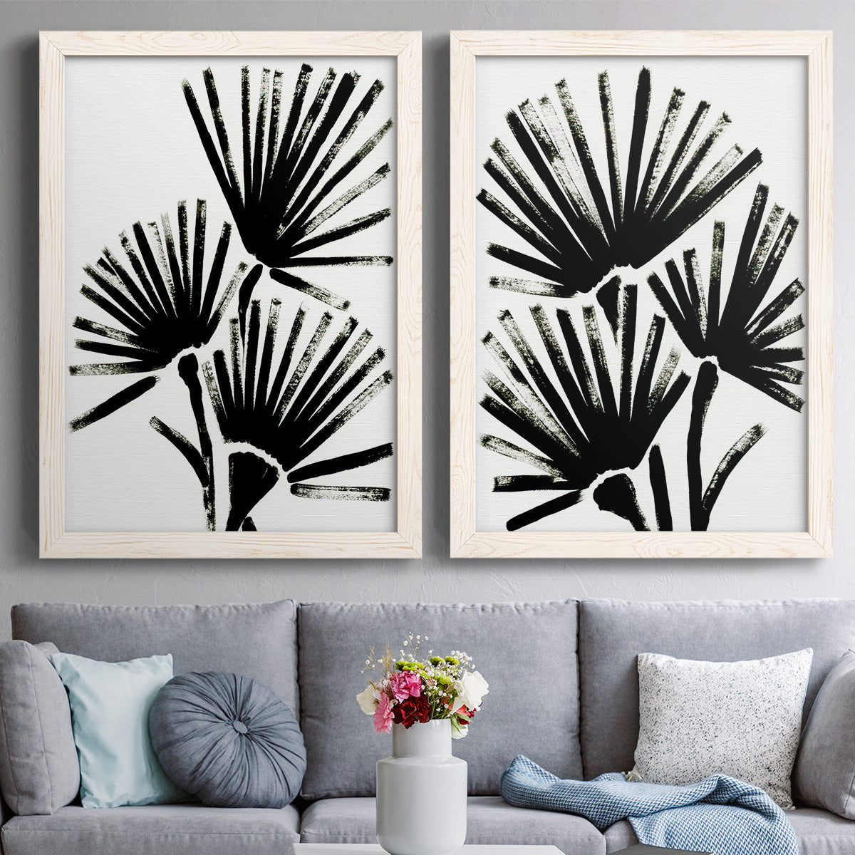 Set of two white barnwood framed canvas prints depicting paired bold monochrome botanical prints with radiating brush marks, repeated leaf-like fans, and white grounds above a sofa in a contemporary living room.