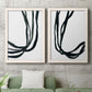 Set of two white barnwood framed canvas prints depicting a coordinated art pair of flowing black ribbon-like lines rendered as minimalist gestural line abstracts above a sofa in a modern living room.
