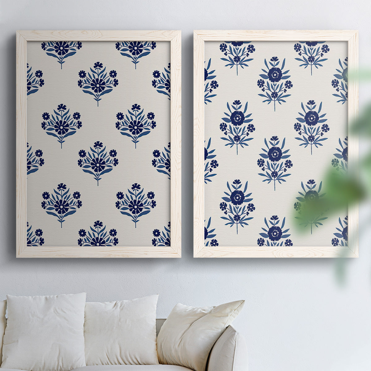 Set of two white barnwood framed canvas prints depicting decorative folk inspired florals rendered in deep blue on soft cream fields above a sofa in a bright living space.