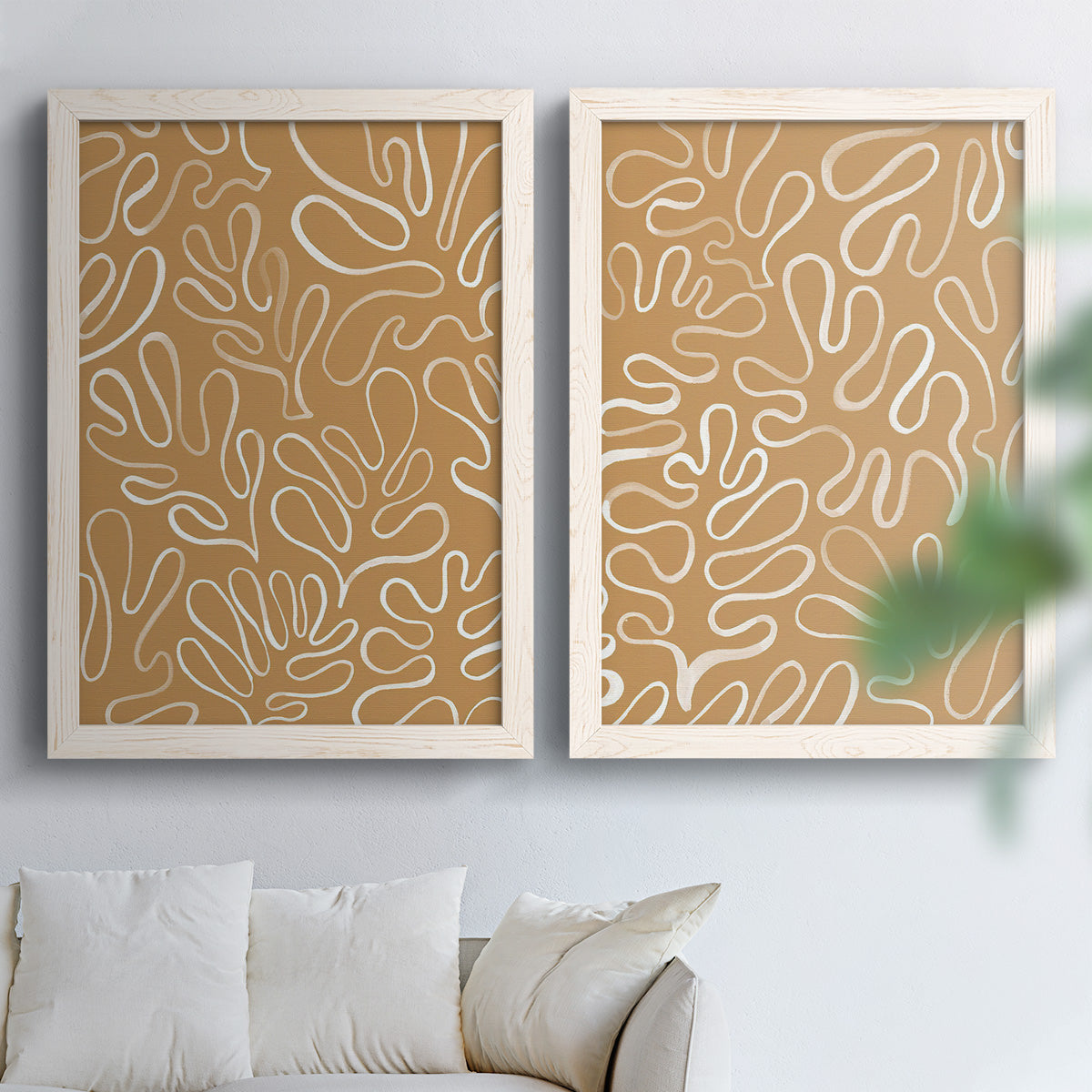 Set of two white barnwood framed canvas prints depicting leaf-like contours, flat warm backgrounds, and repeated flowing shapes arranged across two complementary compositions above a sofa in a bright living space.